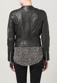 Schwarze Lederjacke mit einer taillierten Passform, die eine glatte Textur, markante Nähte und silberne Reißverschlüsse aufweist. Über einer gemusterten Bluse getragen.