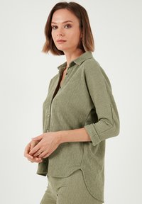 Chemise olive verte à boutons, en tissu texturé et froissé ; dotée d'un col et de manches retroussées ; l'ourlet est légèrement arrondi.
