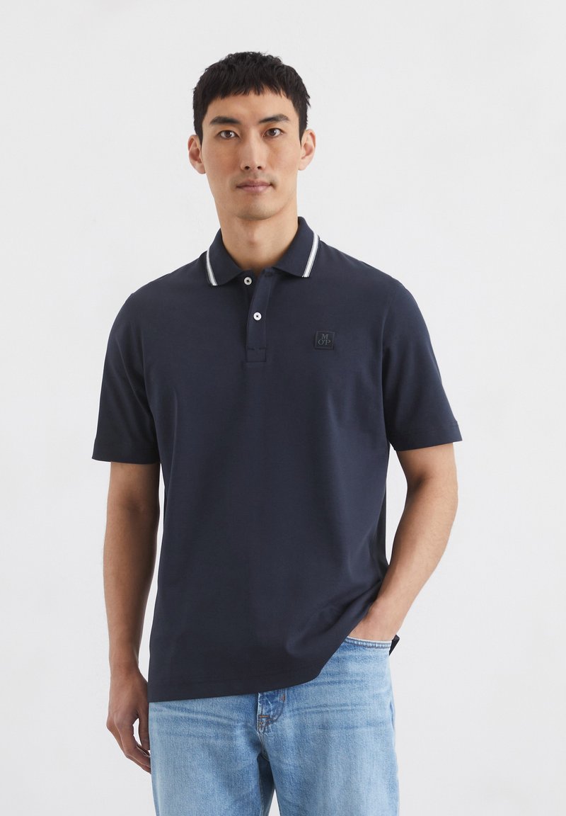 Navy polo shirt with a white striped collar, short sleeves, and a front pocket featuring a black logo. Soft, casual fabric.