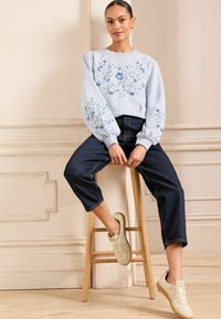 Svetlo modra jakna s cvetličnim vezenjem, napihnjeni rokavi, temno modri jeans in bež superge na lesenem stolu proti nevtralni ozadju.