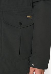 Black jacket with a textured finish, featuring a large flap pocket, a Volcom logo, and snap button closure on the cuff.