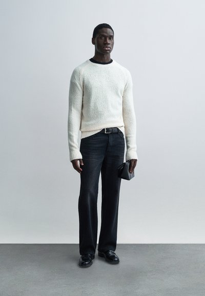 White textured knit sweater, dark denim jeans, black belt, black shoes, holding a small black bag, against a light gray background.