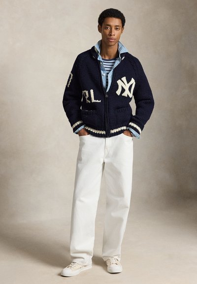 Jeune homme debout les mains dans les poches, portant un pull zippé bleu marine avec des lettres blanches, une chemise en denim clair, un haut rayé, un pantalon blanc et des baskets.