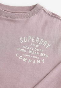 Šviesiai violetinė džemperis su šonkačio apykakle ir išspausdintu baltu tekstu, kuriame rašoma "SUPERDRY JPN HEAVY DUTY WORK-WEAR MFG. COMPANY", išdėstytu išlenktoje formoje.