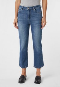 Mittelhohe blaue Denim-Jeans mit geradem Bein, verkürztem Saum, klassischem Fünf-Taschen-Stil und subtiler Ausbleichung. Kombiniert mit schwarzen Slippern.