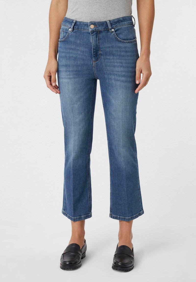 Mittelhohe blaue Denim-Jeans mit geradem Bein, verkürztem Saum, klassischem Fünf-Taschen-Stil und subtiler Ausbleichung. Kombiniert mit schwarzen Slippern.