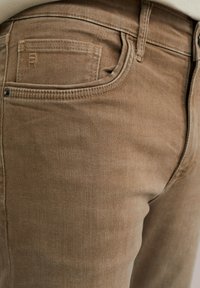 Värikkäät farkut ruskea denim, joissa on teksturoitu pinta. Takana on pieni tasku ja hienovaraisia ompeleita. Vyötärö on hieman kohotettu.
