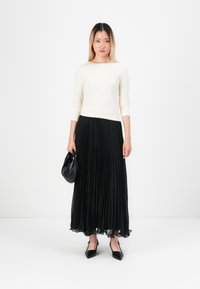Mulher com cabelo até aos ombros, vestindo uma blusa de manga longa em tom creme, uma saia maxi plissada preta e sapatos de bico fino pretos, segurando uma mala preta.