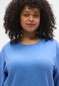 Μπλε πουλόβερ από φλις με στρογγυλή λαιμόκοψη, μακριά μανίκια και υφασμένη υφή. Το μοντέλο φοράει ένα απλό ασημένιο κολιέ με διακριτική λάμψη.