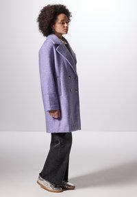 Cappotto doppio petto lilla in tessuto testurizzato, con risvolti oversize, tasche laterali e bottoni coordinati, indossato sopra pantaloni neri a zampa e scarpe argentate.