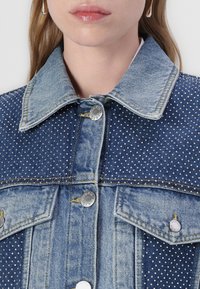 Denim jacket with a collared neckline, featuring a polka dot pattern, silver buttons, and denim texture in light blue shades.