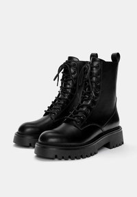 Bottes de combat en cuir noir avec un laçage à l'avant, bout rond et semelles en caoutchouc épaisses. Dotées d'une cheville rembourrée et de languettes à l'arrière.