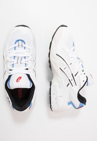 Vita sportskor i vitt med mesh och syntetmaterial, svarta accenter, blåa och röda detaljer, framträdande snörning och texturerade sulor.