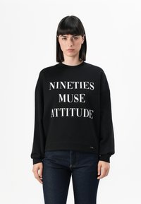 Felpa nera con testo bianco che riporta la scritta "NINETIES MUSE ATTITUDE." Presenta un collo rotondo e maniche lunghe con una vestibilità comoda.