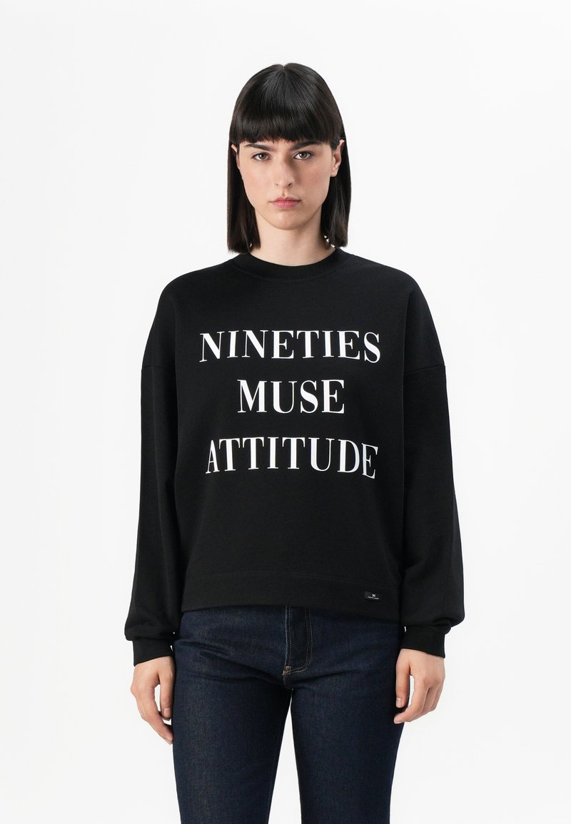 Felpa nera con testo bianco che riporta la scritta "NINETIES MUSE ATTITUDE." Presenta un collo rotondo e maniche lunghe con una vestibilità comoda.