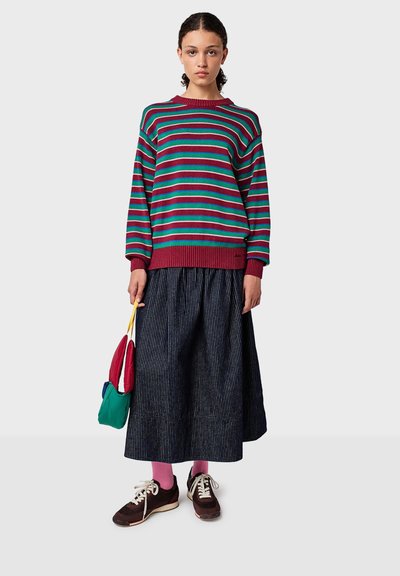 Junge Frau in einem rot, türkis und pink gestreiften Pullover, dunkel gestreiftem Rock, pinken Socken und braunen Sneakern, die eine mehrfarbige kleine Tasche hält.