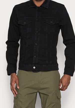 Uomo che indossa una giacca di denim nera aderente con chiusura a bottoni e pantaloni cargo verde oliva, in piedi con le braccia rilassate lungo i fianchi.