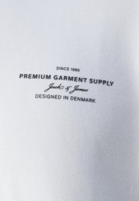Balta medžiaga su lygiu paviršiumi, ant kurios spausdinti juodi tekstai: "PREMIUM GARMENT SUPPLY", "Nuo 1990 metų" ir "Sukurtas Danijoje."