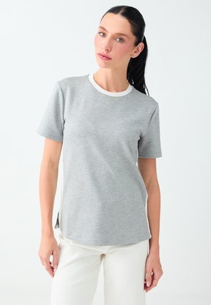 Femme aux cheveux foncés attachés, portant un t-shirt gris à manches courtes avec un bord blanc et un pantalon blanc sur un fond blanc uni.