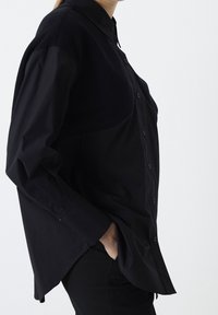 Persona che indossa una camicia nera button-up larga con pannelli superiori testurizzati e maniche lunghe, mano in tasca, su uno sfondo chiaro e semplice.