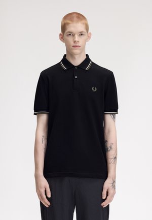 Joven con cabello rojo corto que lleva una camiseta polo negra con cuello y bordes de las mangas a rayas, de pie con expresión neutral sobre fondo blanco.