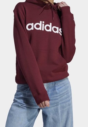 Osoba ubrana w bordową bluzę z kapturem Adidas z białym logo i jasnoniebieskie dżinsy, ręka regulująca sznurek kaptura.