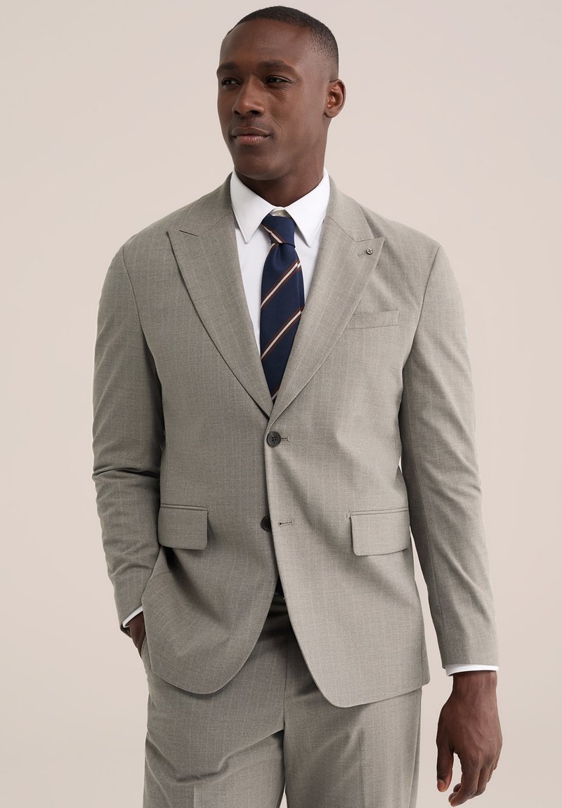 Homme portant un costume gris clair avec une chemise blanche et une cravate rayée bleu marine, debout avec une main dans la poche devant un fond neutre.