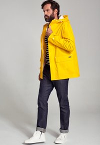 Gul vandtæt jakke med hætte, frontlomme, parret med mørke denimjeans og hvide sneakers. Stribet skjorte synlig under.