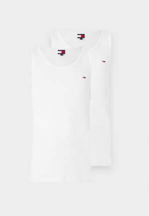 Ung mann iført hvit singlet, blå jeans, hvite joggesko og en olivengrønn caps, stående foran en ensfarget hvit bakgrunn.