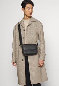 Borsa a tracolla nera con texture, chiusura con zip e tracolla regolabile, indossata sopra un cappotto a quadri beige chiaro e una maglietta beige.