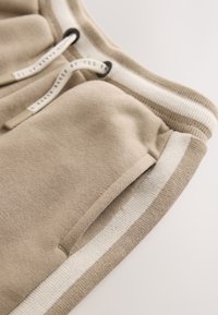 Joggers en coton beige à finition texturée, avec une ceinture à rayures blanches et des cordons de serrage, ainsi qu'une poche latérale fonctionnelle.