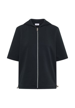 Femme portant un débardeur rose, un sweat à capuche zippé bleu marine à manches courtes, un short gris à cordon et des baskets beiges, debout contre un mur beige.