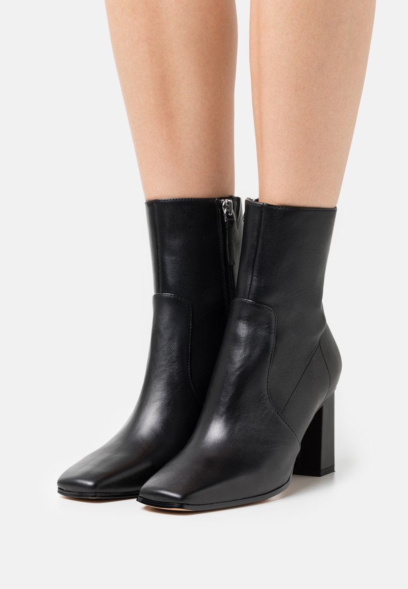 Bottines en cuir noir avec talons carrés et fermetures éclair latérales portées sur des jambes nues sur fond blanc.
