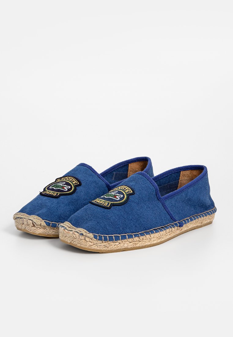 Espadrilles en bleu toile avec une semelle en jute tissée, présentant des patchs brodés sur le dessus et un design à enfiler. Construction robuste.