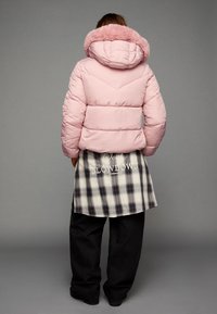 Puhasti kaput s mekim ružičastim slojem i umjetnim krznom na ovratniku. Sadrži prošiveni dizajn i chevron uzorak. Uparen je s dugim karo košuljom i tamnim hlačama.