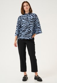 Blusa con stampa zebra nera e blu, colletto alla mandarino, chiusura con bottoni e maniche ampie, abbinata a pantaloni neri su misura e mocassini.