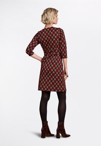 Rotes karierter Kleid mit Dreiviertelärmeln und tailliertem Schnitt. Aus weichem Stoff gefertigt, verfügt es über einen ausgestellten Rock, kombiniert mit glänzenden braunen Stiefeletten.