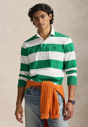 Polo à manches longues rayé vert et blanc avec col, associé à un pull orange texturé noué autour de la taille. Jean en denim visible.