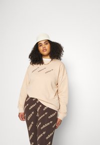 Beige sporta krekls ar brūnu "adidas" logo, brīvs piegriezums. Brūnas bikses ar atkārtotu gaiši brūnu "adidas" logo un lapu rakstu. Līdzenas malas cepure.