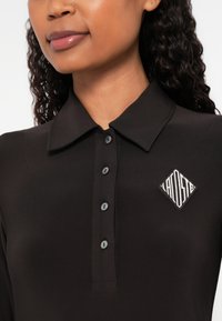 Fekete galléros póló, hegyes gallérral, öt gombbal és gyémánt alakú logóval. A szövet simának és testre szabottnak tűnik.