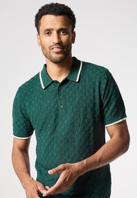 Polo de color verde con textura y patrón a cuadros, cuello y puños en contraste blanco, tapeta de tres botones y mangas cortas.