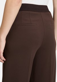Pantalon marron ajusté avec une texture lisse, présentant une taille côtelée et deux fausses poches arrière, vu de côté.