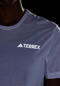Camiseta deportiva ligera de tela gris con una textura suave, que presenta un prominente logo blanco "TERREX" en el pecho.