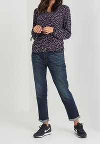 Donna che indossa una blusa floreale a maniche lunghe blu navy, jeans scuri con risvolto e sneakers nere Nike, in piedi contro uno sfondo chiaro e uniforme.