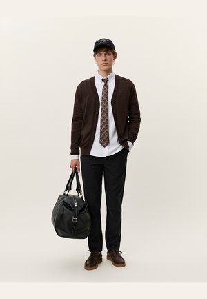 Brun cardigan over en hvid skjorte og mønstret slips, sorte bukser, mørke sko og en sort taske. Modellen har en sort kasket på.