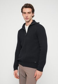 Σκούφο πλεκτό φούτερ σε σκούρο ναυτικό μπλε με φερμουάρ και ριμπ υφή, με κουκούλα, εμπρόσθιο φερμουάρ και διακριτικό λογότυπο στο τελείωμα. Φορεμένο με μπεζ παντελόνι.