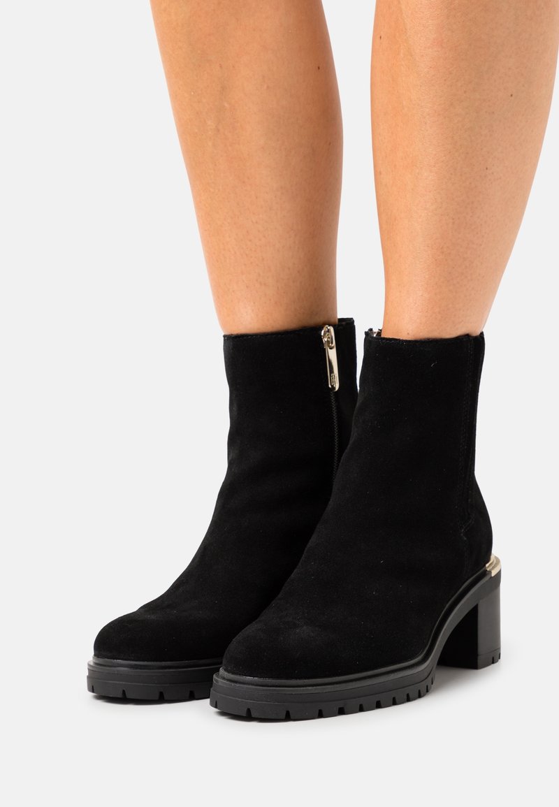 Bottes chevilles en daim noir avec talons épais et fermetures éclair latérales portées sur des jambes nues sur un fond clair uni.