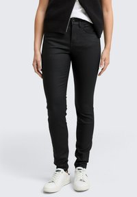 Svarta slim-fit jeans med en slät textur, fem fickor och bälteshällor, kombinerade med vita sneakers. Designen är enkel och mångsidig.
