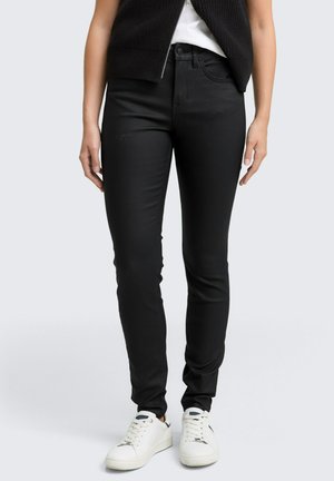 Svarta slim-fit jeans med en slät textur, fem fickor och bälteshällor, kombinerade med vita sneakers. Designen är enkel och mångsidig.