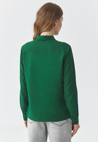 Green long-sleeve shirt made of smooth fabric. Features a collar and button cuffs. Back view shows a tailored fit and clean design.